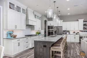 Kitchen featuring white cabinets, custom range hood, a kitchen breakfast bar, a center island with sink, and appliances with stainless steel finishes