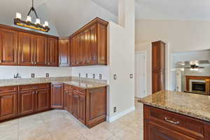 Kitchen with high vaulted ceiling, a tiled fireplace, ceiling fan with notable chandelier, light stone counters, and light tile patterned flooring