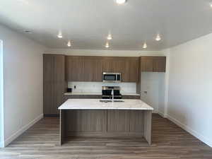 Kitchen featuring a kitchen island with sink, a textured ceiling, stainless steel appliances, light stone counters, and hardwood / wood-style flooring