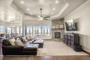 Living room featuring light hardwood / wood-style flooring, ceiling fan with notable chandelier, a raised ceiling, and a stone fireplace