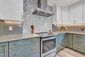 Kitchen with stainless steel electric range, light hardwood / wood-style floors, white cabinetry, wall chimney exhaust hood, and decorative backsplash