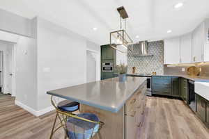 Kitchen featuring light hardwood / wood-style flooring, appliances with stainless steel finishes, hanging light fixtures, white cabinetry, and wall chimney range hood