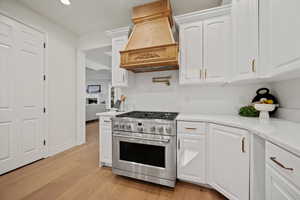 Kitchen with light wood-type flooring, custom exhaust hood, white cabinetry, light stone counters, and high end stove