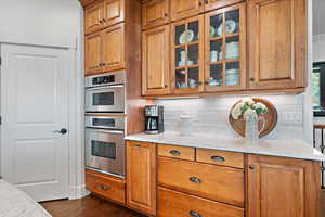 Kitchen featuring dark wood-type flooring, stainless steel double oven, light stone countertops, and decorative backsplash