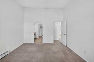 Unfurnished bedroom featuring arched walkways, a high ceiling, a baseboard heating unit, ensuite bath, and light carpet