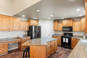 Kitchen with dark wood-type flooring, black appliances, a kitchen island, and light stone counters