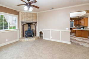 Unfurnished living room with ceiling fan, light carpet, ornamental molding, and a wood stove