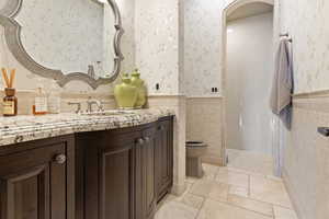 Full bathroom with tile walls, vanity, toilet, wainscoting, and wallpapered walls