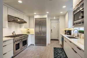 Kitchen with built in appliances, extractor fan, sink, white cabinetry, and light stone counters