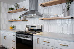 Kitchen with stainless steel range with gas stovetop, decorative backsplash, white cabinetry, light stone counters, and wall chimney range hood