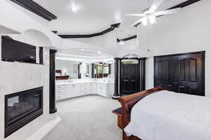 Carpeted bedroom with ceiling fan, ensuite bath, decorative columns, and a fireplace