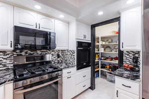 Kitchen with dark stone countertops, black appliances, white cabinetry, and backsplash