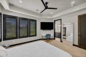 Carpeted bedroom featuring a tray ceiling, ensuite bathroom, and ceiling fan