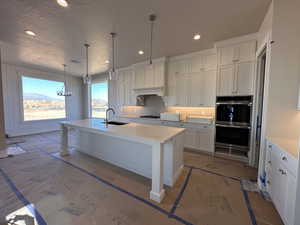 Kitchen with sink, hanging light fixtures, an island with sink, white cabinets, and double oven