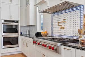 Kitchen with exhaust hood, decorative backsplash, light wood-type flooring, appliances with stainless steel finishes, and white cabinetry