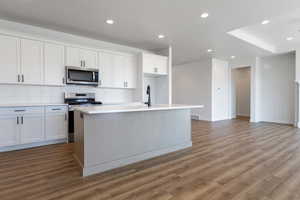 Kitchen with appliances with stainless steel finishes, dark wood-type flooring, white cabinets, backsplash, and a center island with sink