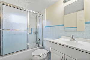 Full bathroom with toilet, tile walls, vanity, bath / shower combo with glass door, and a textured ceiling