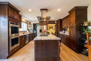 Kitchen with appliances with stainless steel finishes, light hardwood / wood-style floors, a center island, light stone counters, and dark brown cabinetry, hood vents to outside