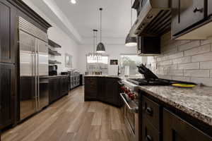 Kitchen with range hood, hanging light fixtures, backsplash, light stone countertops, and premium appliances