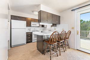 Kitchen featuring white appliances, dark brown cabinets, a kitchen bar, kitchen peninsula, and light colored carpet