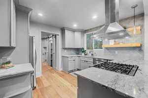 Kitchen featuring decorative light fixtures, tasteful backsplash, island range hood, stainless steel appliances, and light wood-type flooring