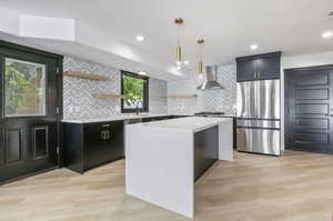 Kitchen featuring a center island, stainless steel appliances, light hardwood / wood-style floors, wall chimney range hood, and pendant lighting