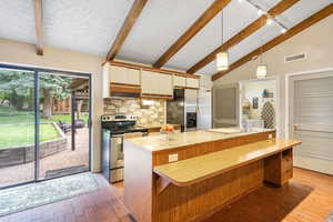 Kitchen featuring decorative light fixtures, a center island with sink, stainless steel appliances, white cabinetry, and lofted ceiling with beams