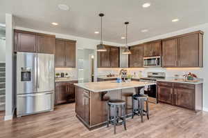 Kitchen with decorative light fixtures, appliances with stainless steel finishes, light hardwood / wood-style floors, an island with sink, and dark brown cabinetry