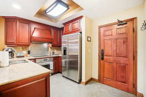 Kitchen with light stone counters, a tray ceiling, sink, appliances with stainless steel finishes, and premium range hood