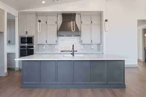 Kitchen with gray cabinetry, lofted ceiling, a kitchen island with sink, custom range hood, and appliances with stainless steel finishes