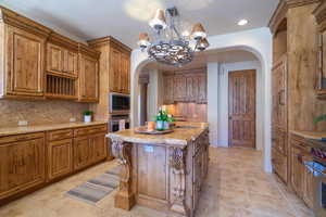 Kitchen with appliances with stainless steel finishes, decorative backsplash, a kitchen island, light stone countertops, and a notable chandelier