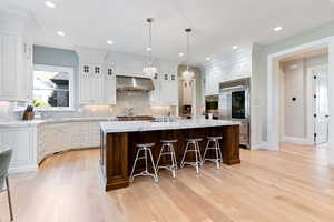 Kitchen featuring tasteful backsplash, white cabinetry, wall chimney range hood, white oak floors, and a center island with sink