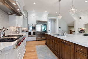 Kitchen with wall chimney exhaust hood, white cabinetry, appliances with stainless steel finishes, a notable chandelier, and white oak floors