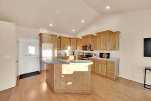 Kitchen with lofted ceiling, a kitchen island with sink, light brown cabinetry, appliances with stainless steel finishes, and light hardwood / wood-style floors