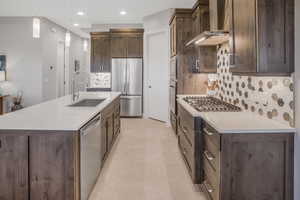 Kitchen with sink, wall chimney exhaust hood, stainless steel appliances, a center island with sink, and decorative light fixtures