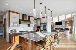 Kitchen with a large island with sink, white cabinetry, a stone fireplace, wall chimney exhaust hood, and high end appliances