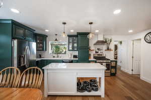 Kitchen featuring light wood-type flooring, a center island, wall chimney range hood, stainless steel appliances, and decorative light fixtures