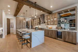 Kitchen with wine cooler, hanging light fixtures, beam ceiling, light hardwood / wood-style flooring, and wall chimney range hood