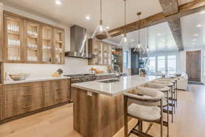 Kitchen with light hardwood / wood-style floors, beamed ceiling, sink, wall chimney range hood, and a spacious island