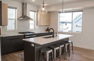 Kitchen with hanging light fixtures, a center island with sink, sink, and wall chimney range hood