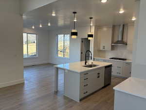 Kitchen featuring a kitchen island with sink, white cabinets, wall chimney range hood, sink, and hanging light fixtures