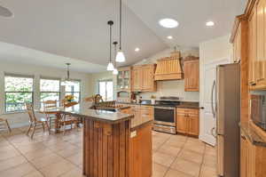 Kitchen featuring custom exhaust hood, lofted ceiling, an island with sink, light tile patterned floors, and appliances with stainless steel finishes