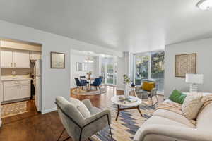 Living room with dark laminate floors and a chandelier opens to the kitchen. Large windows.