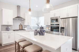 Kitchen with a kitchen island, wall chimney range hood, white cabinetry, stainless steel appliances, and decorative backsplash