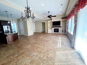 Unfurnished living room with ceiling fan with notable chandelier, a fireplace surrounded by bookshelves and storage, a tray ceiling, and plenty of natural light