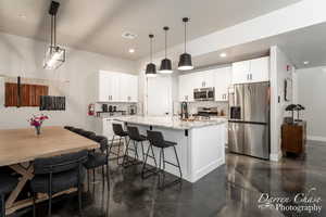 Kitchen with white cabinets, stainless steel appliances, a center island with sink, decorative light fixtures, and light stone countertops