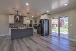 Kitchen with pendant lighting, light hardwood / wood-style floors, white cabinetry, wall chimney range hood, and stainless steel fridge