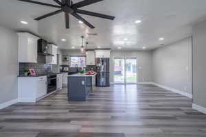 Kitchen with wall chimney exhaust hood, a kitchen island, stainless steel appliances, ceiling fan, and decorative light fixtures