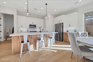 Kitchen with light hardwood / wood-style floors, white cabinets, stainless steel appliances, a center island with sink, and decorative light fixtures