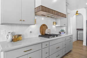 Kitchen with gray cabinets, backsplash, custom exhaust hood, stainless steel gas cooktop, and light wood-type flooring